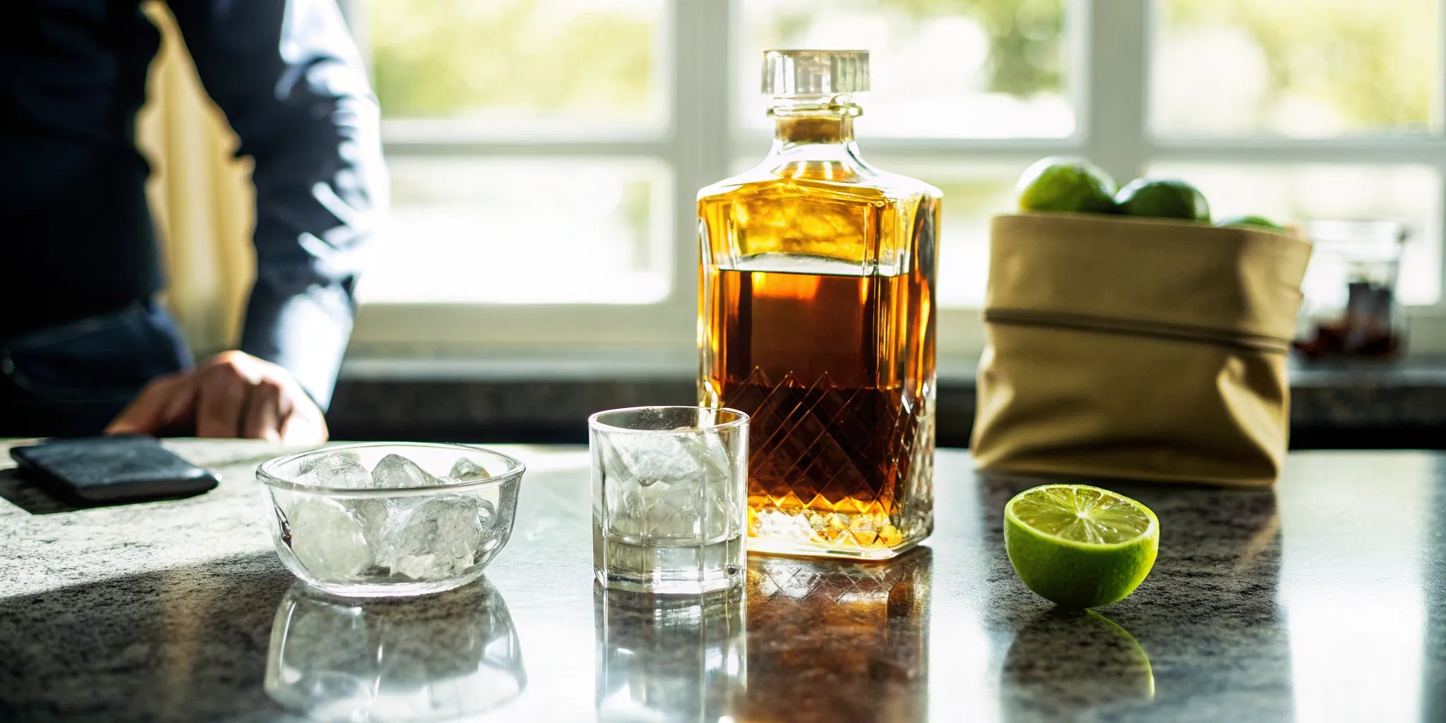 A tequila home delivery setup with a decanter, glass, and fresh limes on a kitchen counter.