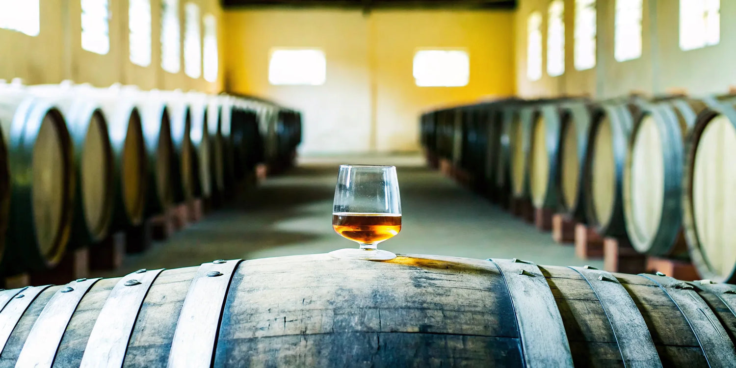 Amber añejo tequila in a glass on an oak barrel in a Jalisco aging warehouse
