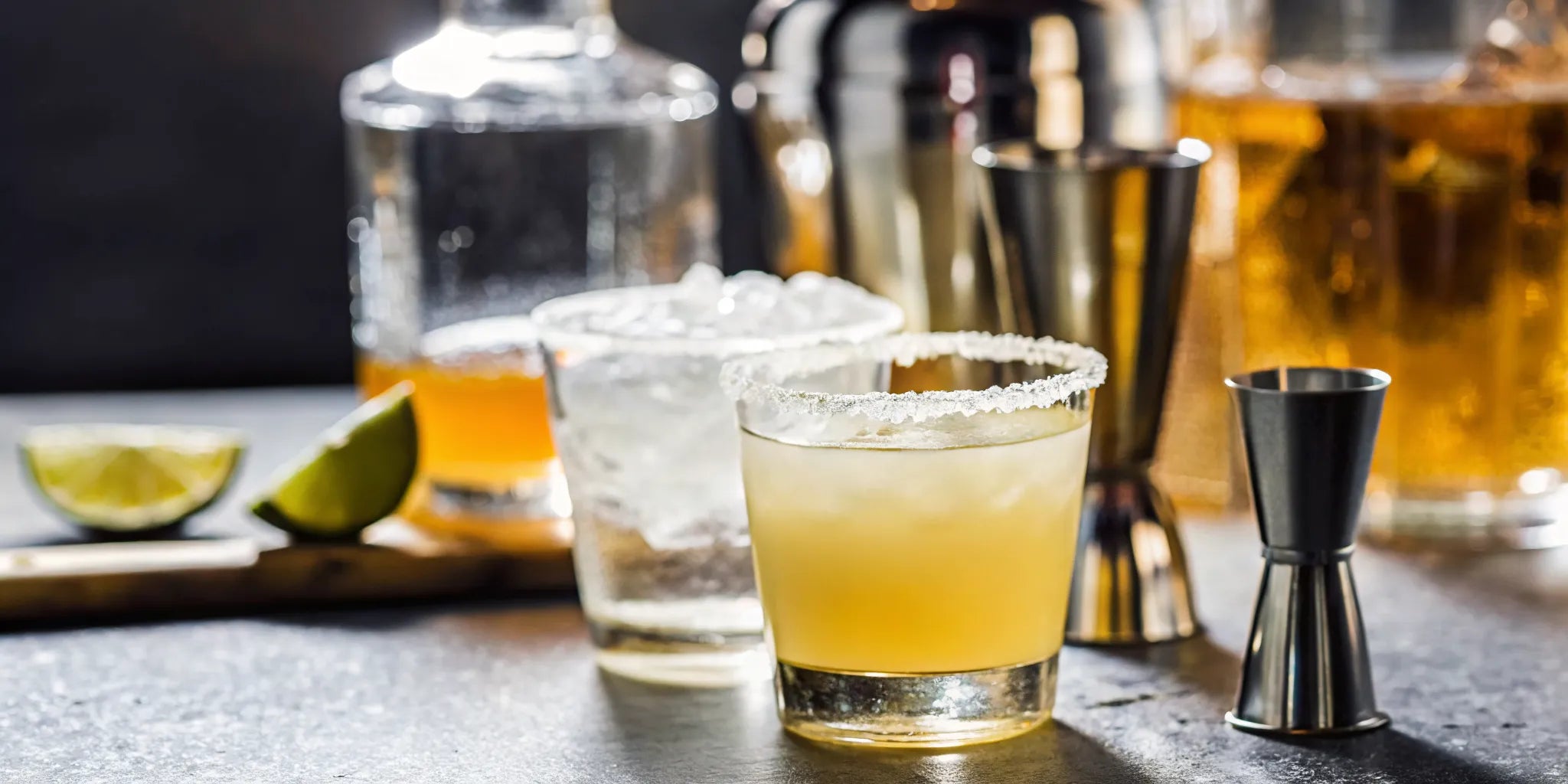 Home bartender shaking a classic margarita beside a bar cart lined with tequila bottles and fresh limes