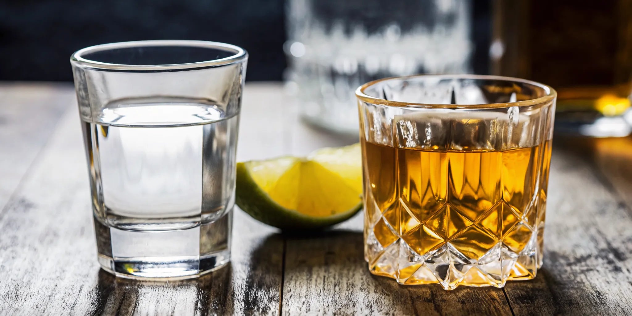 Two glasses side by side on a rustic wooden bar, one filled with clear blanco tequila and one with amber whiskey, warm tasting lighting